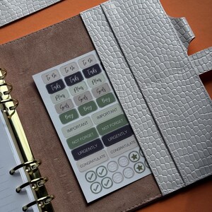 May include: A light gray faux leather planner with a textured, crocodile-like pattern. Inside, a sheet of stickers with task-related labels like "To Do," "Tasks," and "Goals" is visible. The planner has gold-colored metal rings.