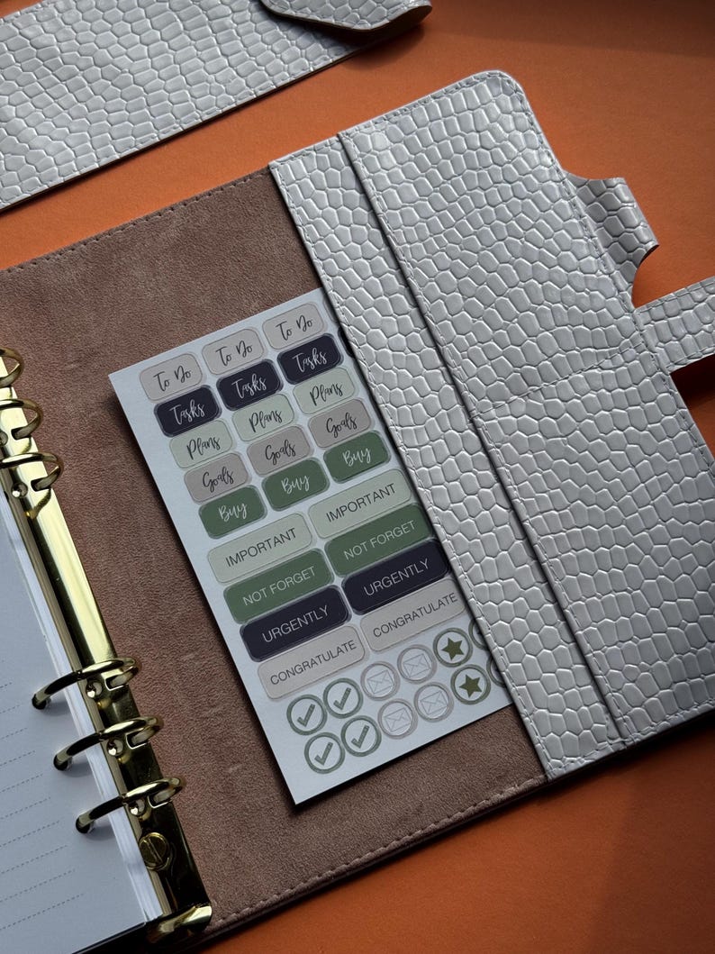May include: A light gray faux leather planner with a textured, crocodile-like pattern. Inside, a sheet of stickers with task-related labels like "To Do," "Tasks," and "Goals" is visible. The planner has gold-colored metal rings.