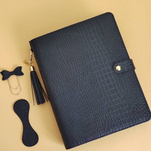 Op de afbeelding: Een zwarte leren planner met een krokodilpatroon, een gouden kwast, een gouden strikclip en een gouden paperclip.