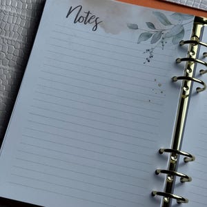 May include: Open planner with lined pages and the word "Notes" in script. The top right corner features a watercolor design of leaves and gold flecks. The planner has gold rings and a textured, light gray cover.