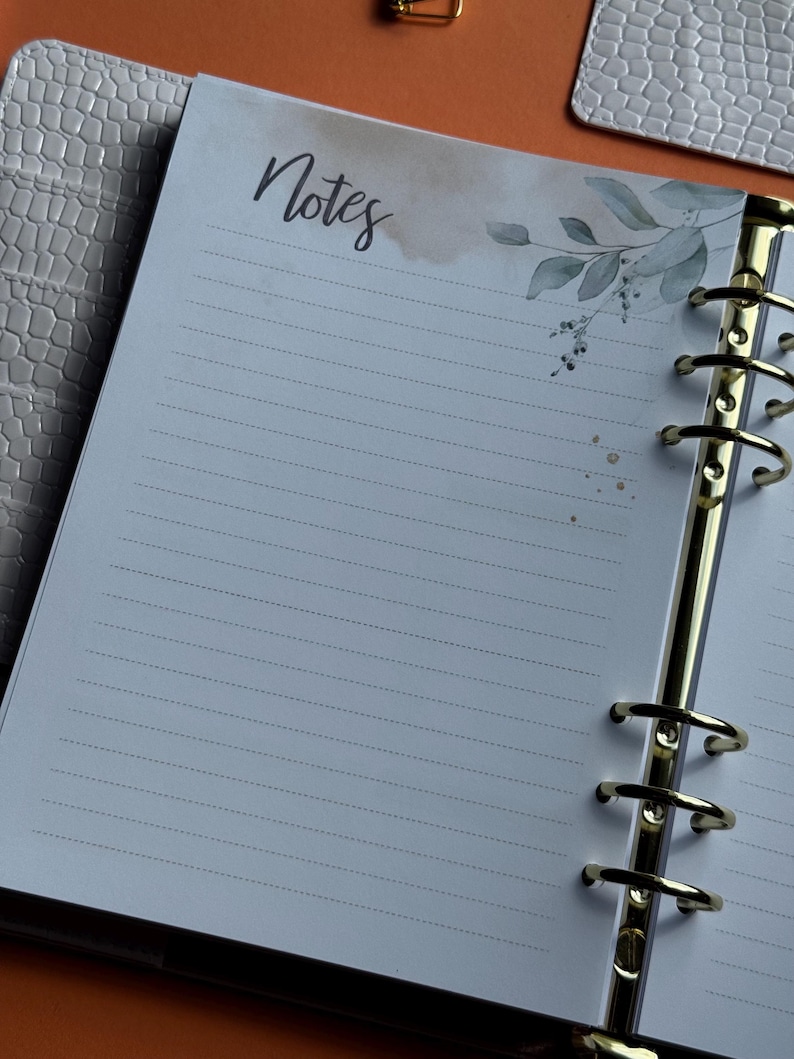 May include: Open planner with lined pages and the word "Notes" in script. The top right corner features a watercolor design of leaves and gold flecks. The planner has gold rings and a textured, light gray cover.