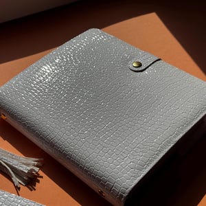 May include: A light gray, textured planner with a reptile-like pattern. The planner has a snap closure with a gold-colored button. A matching tassel and a small accessory are visible in the image. The planner is on an orange surface.