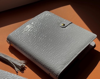 May include: A light gray, textured planner with a reptile-like pattern. The planner has a snap closure with a gold-colored button. A matching tassel and a small accessory are visible in the image. The planner is on an orange surface.