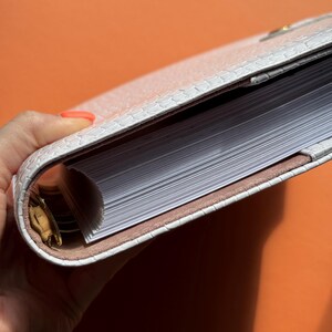 May include: A white, textured planner with a reptile-like pattern. The planner is open, revealing a stack of white pages. The interior lining is a light brown color. The planner is held against an orange background.