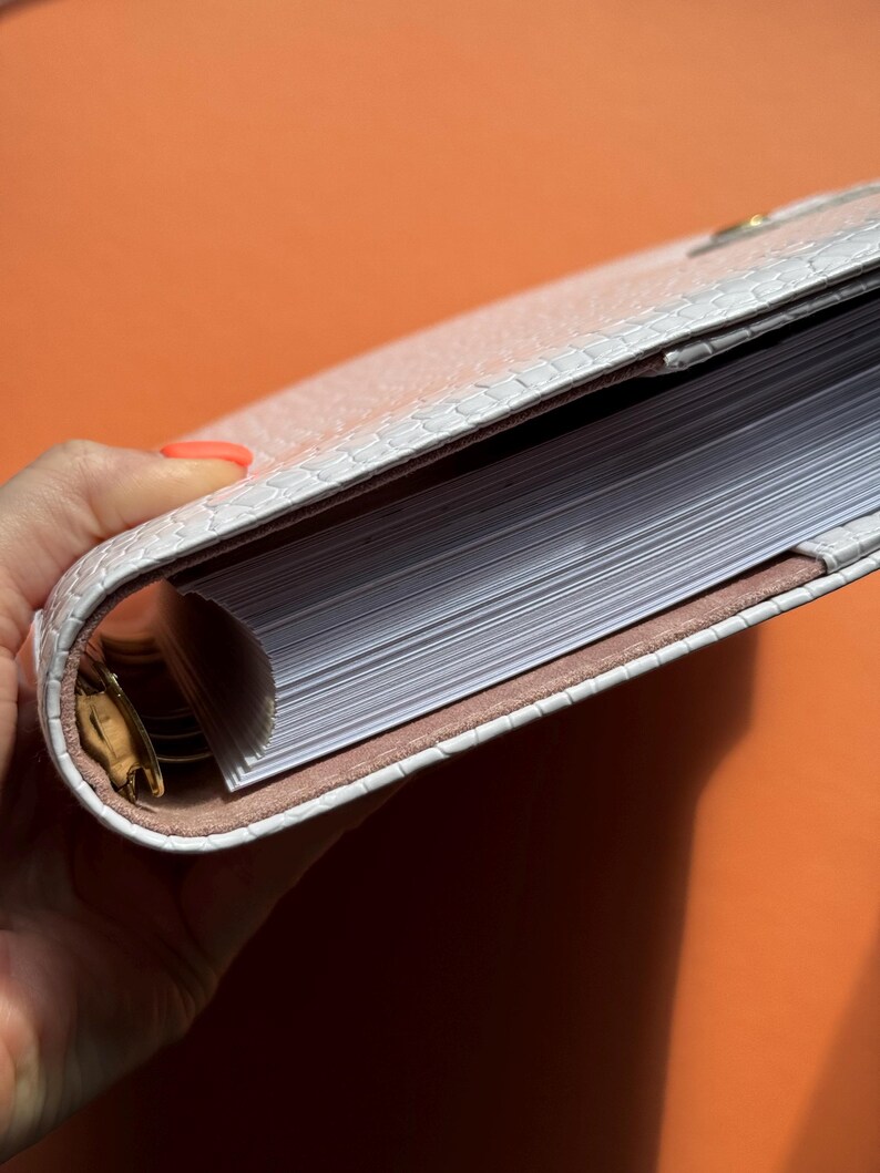 May include: A white, textured planner with a reptile-like pattern. The planner is open, revealing a stack of white pages. The interior lining is a light brown color. The planner is held against an orange background.