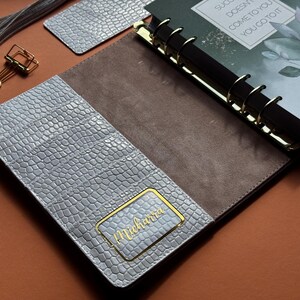May include: Open planner with a gray, textured cover and a brown interior. The planner has gold accents and a gold-colored binder clip. The cover features a gold nameplate that reads "Michara". The planner is open to a page with the text "Success doesn't come to you, you go to it."