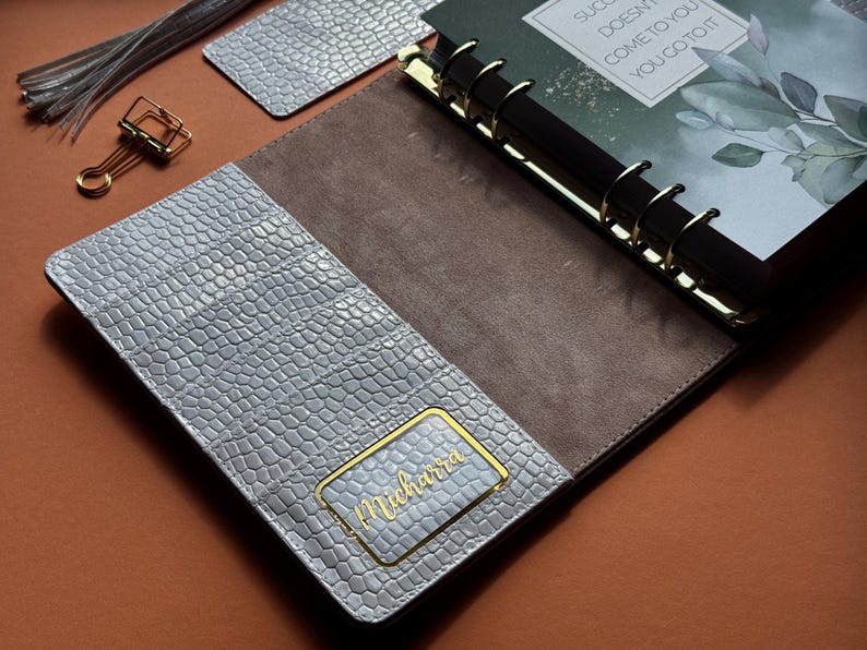 May include: Open planner with a gray, textured cover and a brown interior. The planner has gold accents and a gold-colored binder clip. The cover features a gold nameplate that reads "Michara". The planner is open to a page with the text "Success doesn't come to you, you go to it."