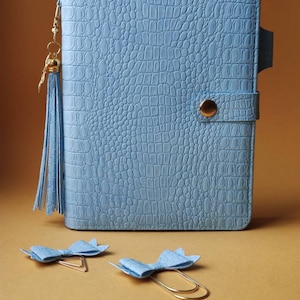 May include: A light blue faux leather planner with a gold tassel and a gold button closure. Two light blue bow-shaped paper clips are on a brown surface.