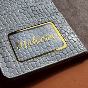 May include: A light blue, textured wallet with a gold-framed clear window. The name "Micharra" is written in gold script within the window. The interior is a contrasting brown suede-like material.