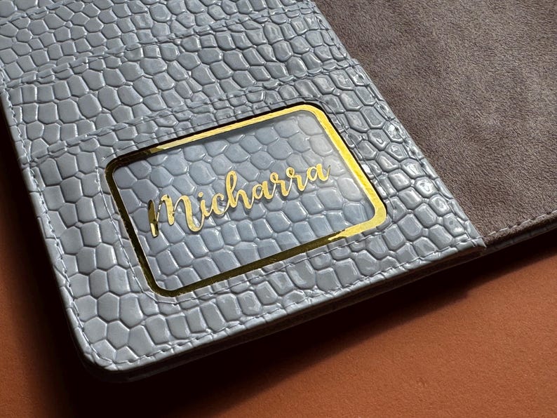 May include: A light blue, textured wallet with a gold-framed clear window. The name "Micharra" is written in gold script within the window. The interior is a contrasting brown suede-like material.