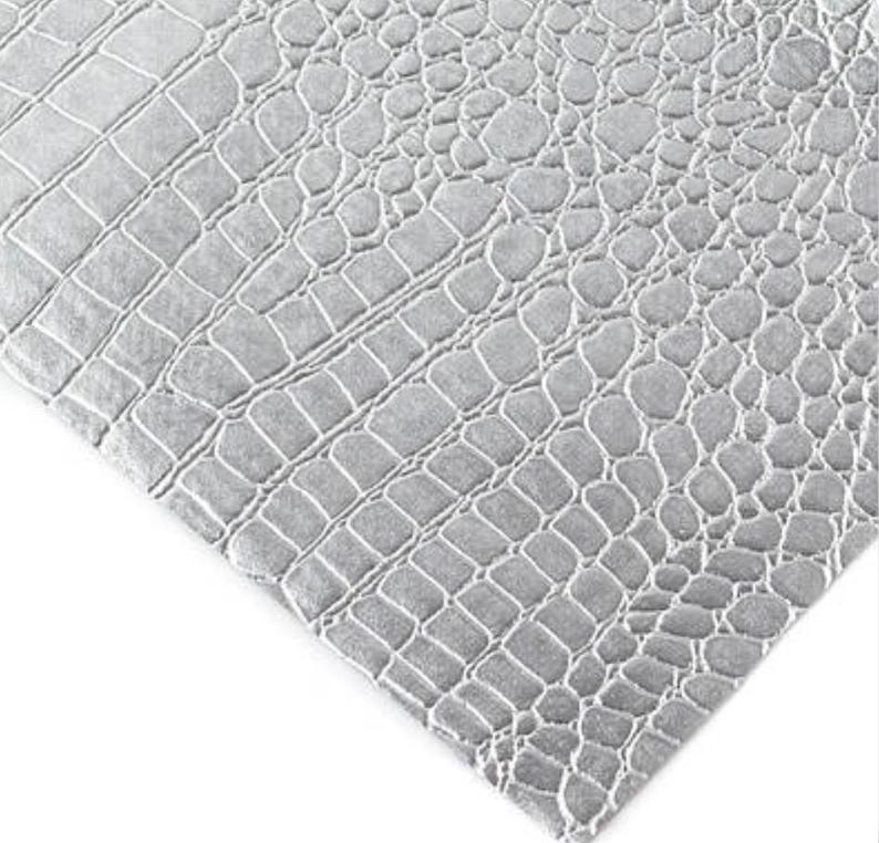 May include: Close-up of a gray, textured material with a crocodile skin pattern. The pattern is made up of irregular, interconnected shapes, creating a realistic reptile skin effect. The material is a light gray color.