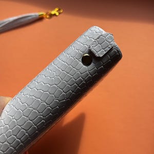 May include: A gray, textured case with a reptile-like pattern. The case is closed with a gold-colored snap. A matching gray tassel with gold hardware is visible in the background.