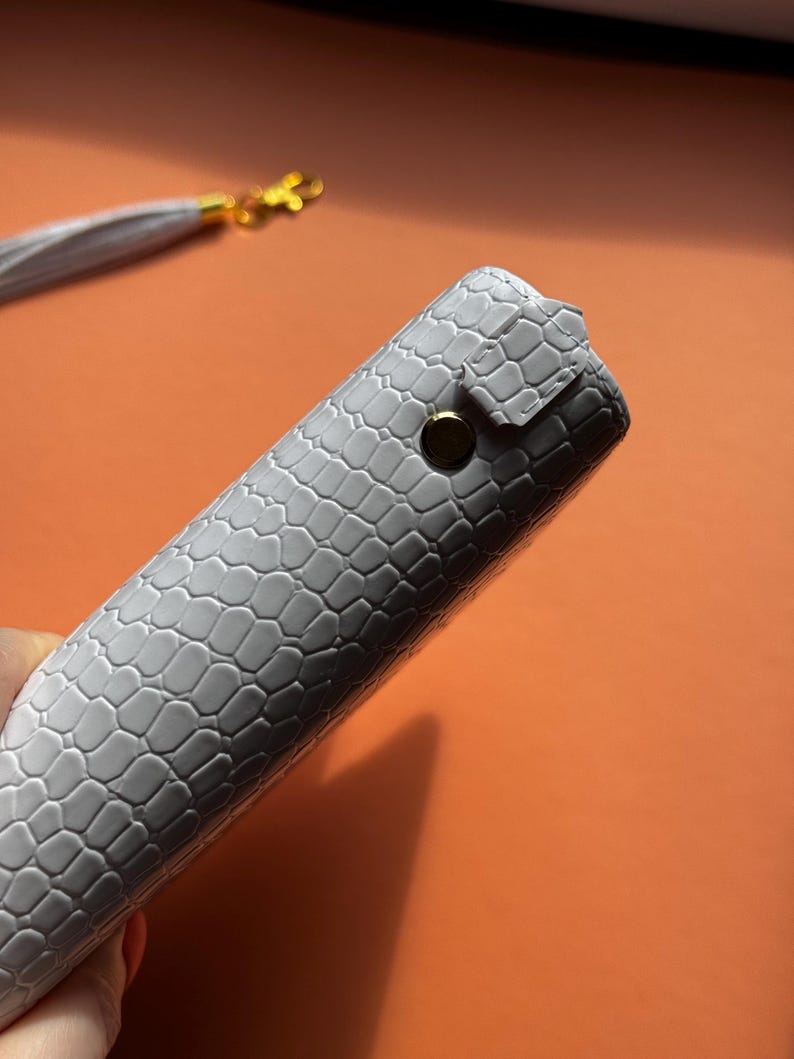 May include: A gray, textured case with a reptile-like pattern. The case is closed with a gold-colored snap. A matching gray tassel with gold hardware is visible in the background.
