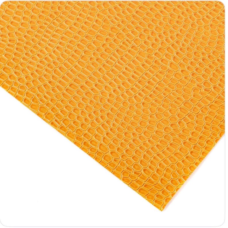 May include: Close-up of a sheet of textured, golden-yellow material. The surface has a repeating pattern of small, interconnected shapes, resembling a honeycomb or reptile skin. The material is angled, with a white background.