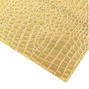May include: Close-up of a gold-colored material with a textured, crocodile-skin pattern. The surface has a repeating design of rounded and rectangular shapes, creating a realistic reptile skin effect. The material is angled, showing the texture and color variations.