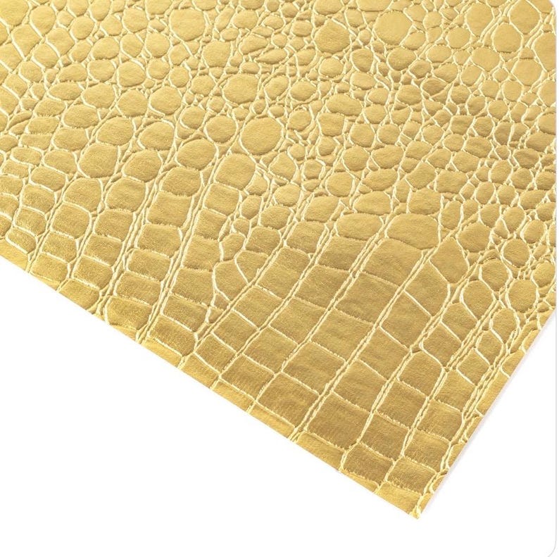May include: Close-up of a gold-colored material with a textured, crocodile-skin pattern. The surface has a repeating design of rounded and rectangular shapes, creating a realistic reptile skin effect. The material is angled, showing the texture and color variations.