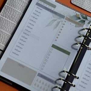 May include: Open planner with a daily layout. The pages feature sections for "Today's Plans," "Today's Goals," and "To Do List." The design includes watercolor-style botanical accents and a water intake tracker. The planner is bound with gold rings.