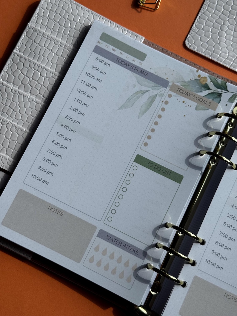 May include: Open planner with a daily layout. The pages feature sections for "Today's Plans," "Today's Goals," and "To Do List." The design includes watercolor-style botanical accents and a water intake tracker. The planner is bound with gold rings.