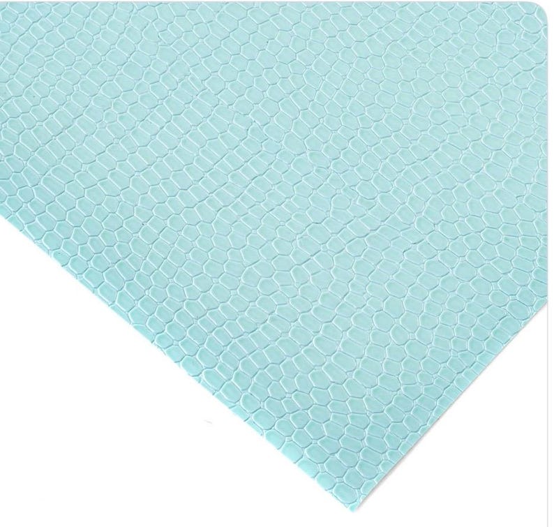 May include: A light blue, textured sheet with a reptile scale pattern. The material has a repeating hexagonal design, creating a textured surface. The sheet is angled, showing the pattern in detail.