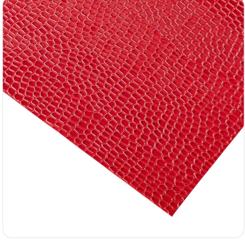 May include: A sheet of red faux leather with a textured, honeycomb-like pattern. The material has a glossy finish and is angled across a white background. The close-up view highlights the detailed texture and vibrant color.