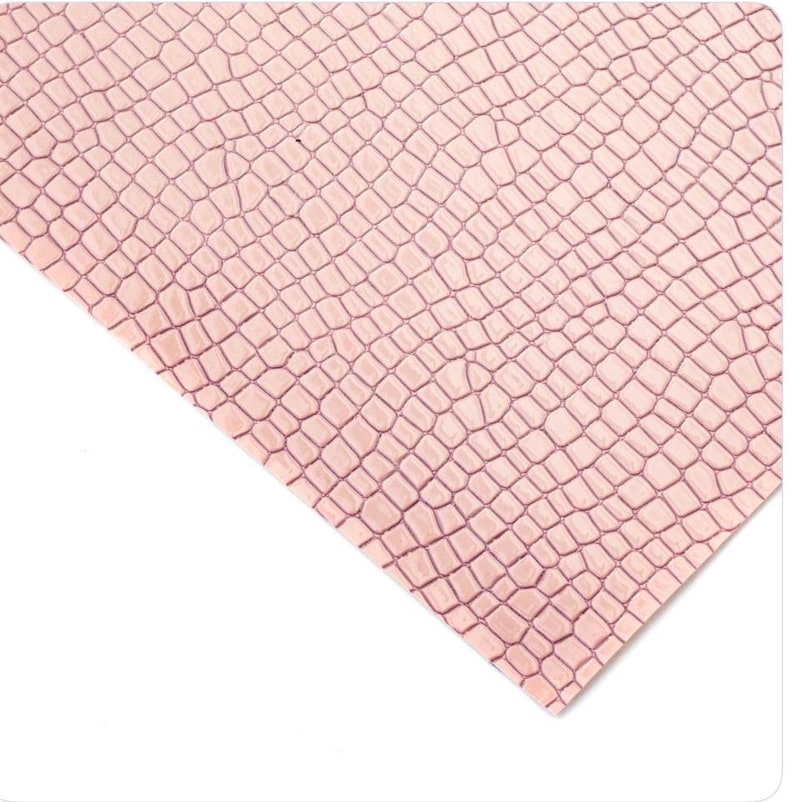 May include: A close-up of a sheet of pink faux leather with a crocodile skin texture. The pattern consists of small, irregular, interconnected shapes, creating a realistic reptile skin effect. The material is angled against a white background.