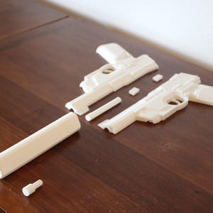 May include: Off-white, disassembled toy gun components arranged on a dark wood surface. The parts include the gun's frame, barrel, and smaller pieces, suggesting a model kit or a disassembled toy.