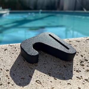 Puede incluir: Un objeto negro texturizado con forma de gancho y una ranura diagonal. El objeto está sobre una superficie clara y texturizada, con una piscina en el fondo.