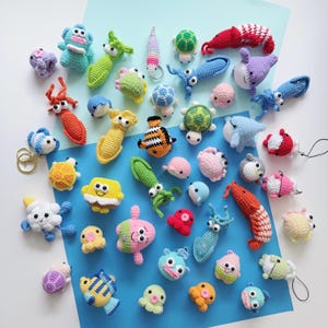 May include: A collection of colorful, handmade crochet sea creatures, including fish, turtles, and octopuses. The toys are in various sizes and colors, with details like googly eyes and keychains. The background is a light blue and white surface.