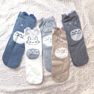 May include: Five pairs of knee-high socks in various colors: blue, white, gray, and tan. Each sock features a cartoon character face and a circular design. The socks have small, fluffy details at the top.