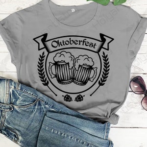 Puede incluir: Una camiseta gris con un dise&ntilde;o gr&aacute;fico negro de dos jarras de cerveza con espuma en la parte superior. El gr&aacute;fico est&aacute; rodeado de una pancarta que dice "Oktoberfest" y una corona de espigas de trigo.