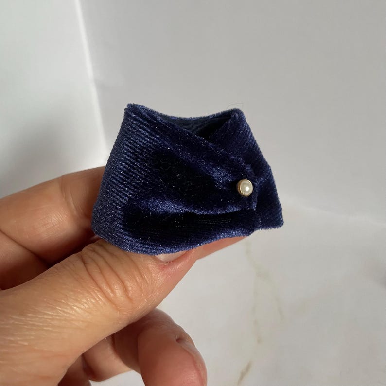 May include: A miniature dark navy blue velvet collar accessory featuring a soft, ribbed texture and a single small, round white faux pearl button detail centered on the front overlap. The fabric has a subtle sheen characteristic of velvet.