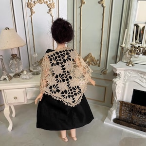 May include: A doll wearing a black dress with a beige crocheted shawl. The doll has dark brown hair and is standing in a room with a fireplace and a table.