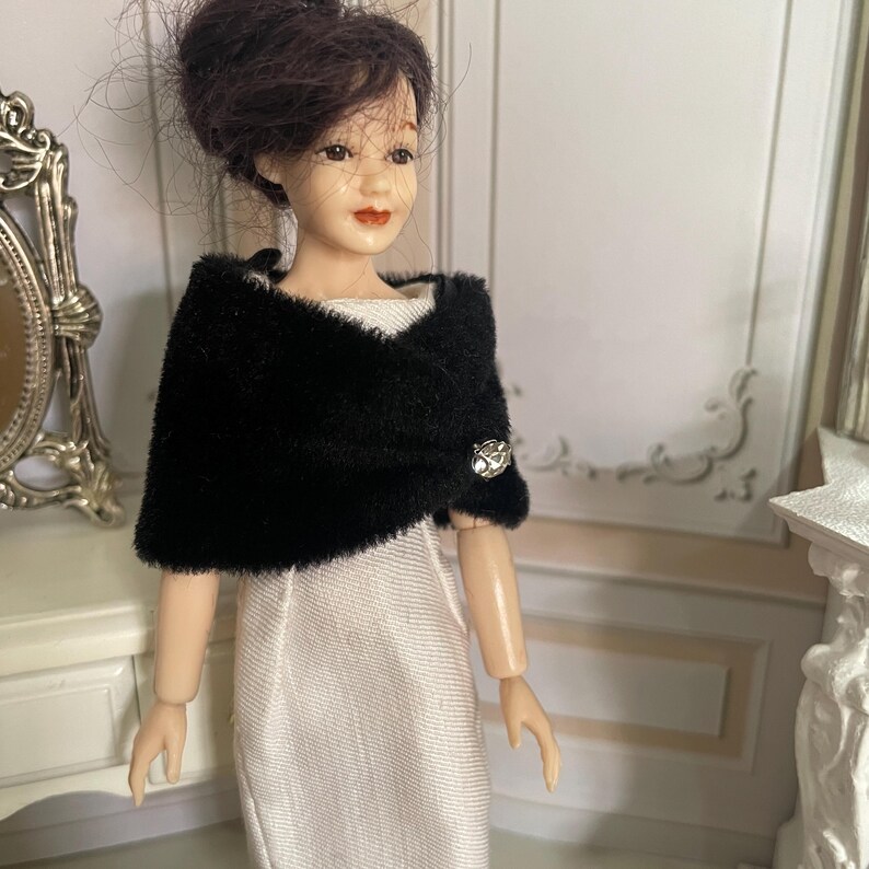 May include: A doll with dark hair styled in an updo, wearing a white sleeveless dress and a black faux fur bolero wrap. The bolero is secured at the front with a single round, faceted silver-toned button featuring a clear central stone.