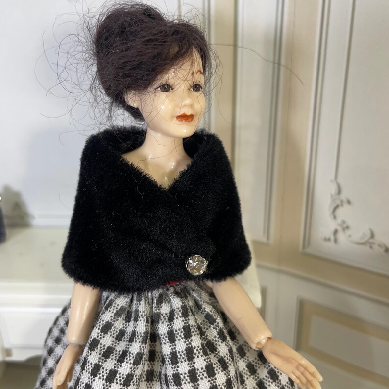 May include: A doll with dark brown hair styled in an updo, wearing a black faux fur bolero with a single clear decorative button closure over a black and white checkered skirt. The doll has painted facial features including red lips and brown eyes.