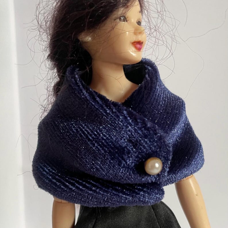 May include: A doll with dark hair wearing a dark blue textured wrap or shawl featuring a subtle shimmer. The garment is secured at the front with a single round, off-white pearl-like button. The doll also wears small white stud earrings.