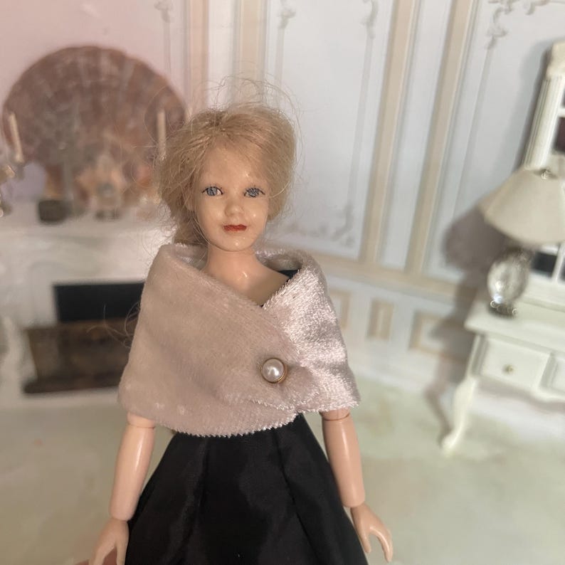 May include: A doll with blonde hair styled in an updo, wearing a light beige velvet shawl fastened with a single pearl-like button over a black dress. The doll has blue eyes and painted facial features, with the shawl draped over the shoulders.