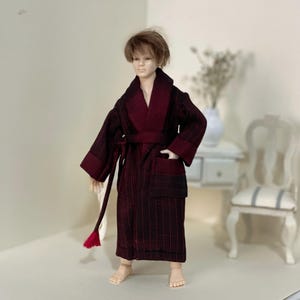 May include: A burgundy and black striped doll-sized robe with a shawl collar, belt, and pockets. The robe has a matching belt and a red tassel. The doll is standing in a room with a white chair and a small table.