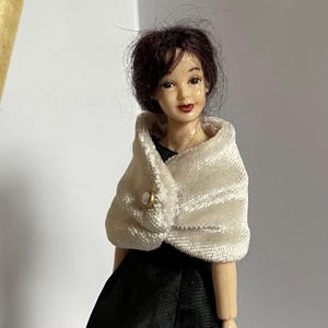 May include: A doll with dark brown hair, painted facial features, and red lips. The doll wears a black sleeveless dress and a white, textured, velvet-like shawl draped over the shoulders, secured at the front with a single round, off-white button.