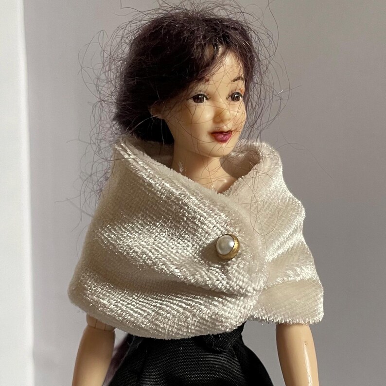May include: A doll with dark brown hair, wearing a cream-colored textured wrap or shawl fastened with a single round white pearl-like button with a gold-toned rim. The doll has a painted face with dark eyes and red lips, and is dressed in a black garment.