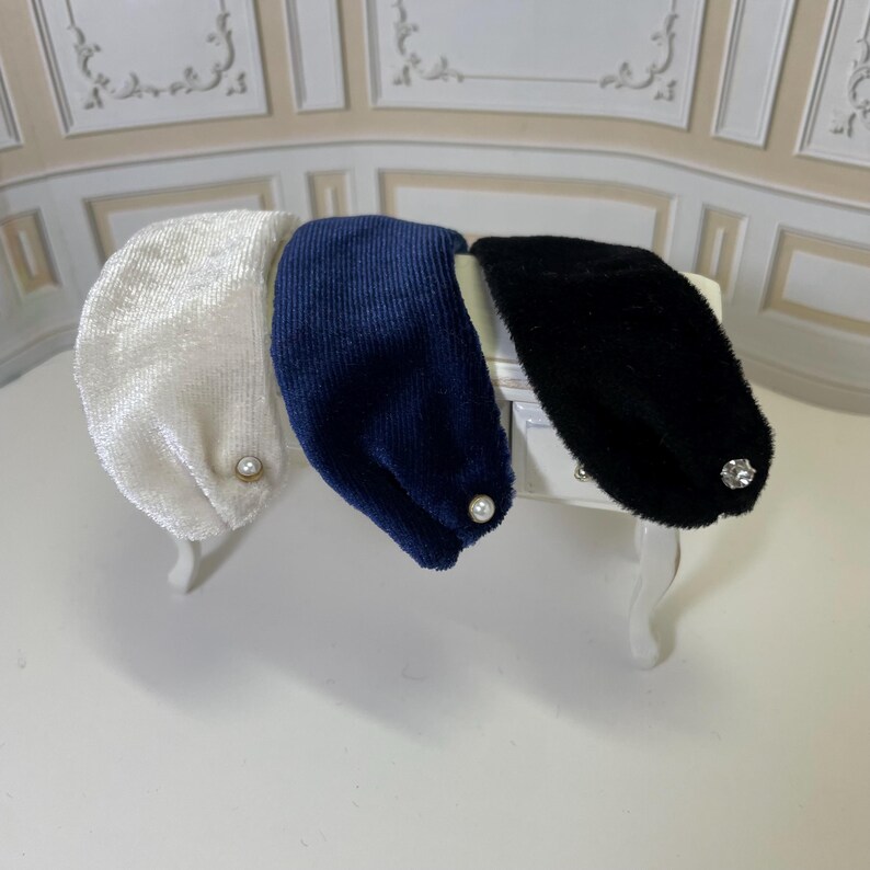 May include: Three wide headbands in white, navy blue, and black. The white headband has a textured finish with a small pearl accent. The navy blue headband features a ribbed corduroy texture with a pearl accent. The black headband is fuzzy with a clear crystal-like embellishment.