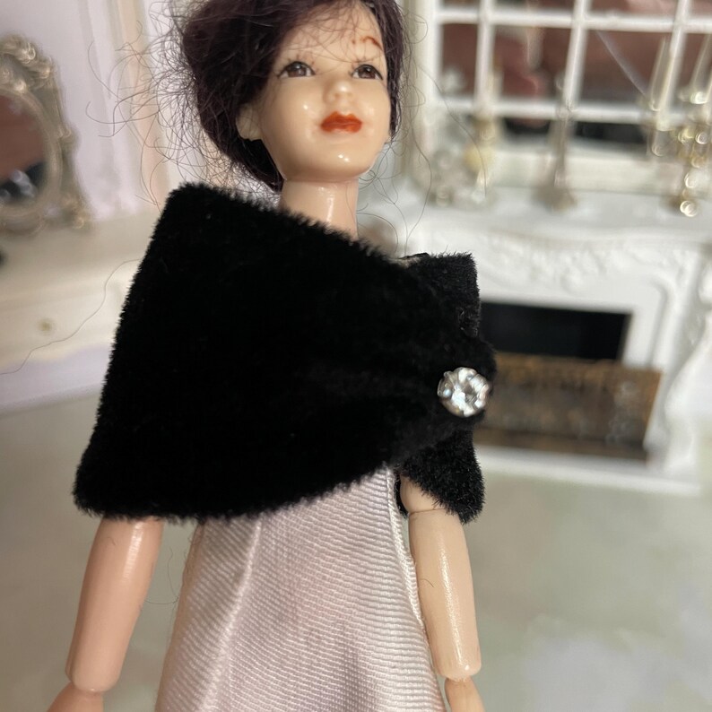 May include: A doll with dark hair wearing a black velvet-textured shawl over a light-colored dress. The shawl is secured at the front with a single round, clear, faceted rhinestone button that reflects light, adding a decorative detail to the garment.