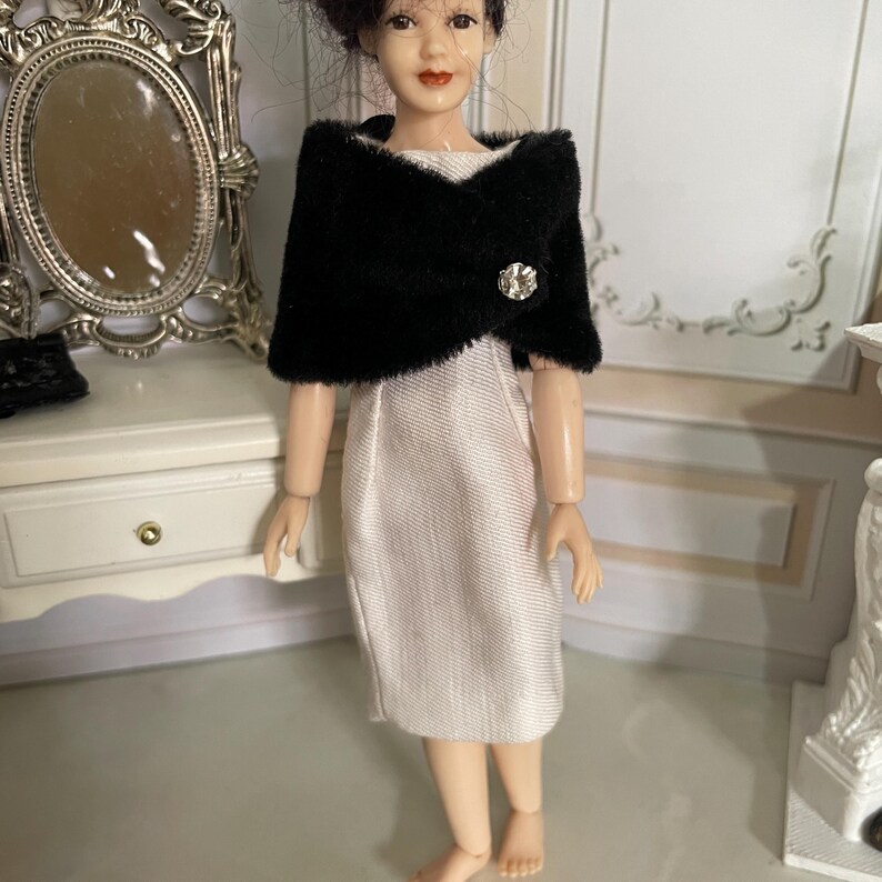 May include: A doll with dark hair wearing a sleeveless white sheath dress and a black faux fur bolero jacket. The bolero features a single clear rhinestone button closure at the front. The doll has a fair complexion and painted facial features.