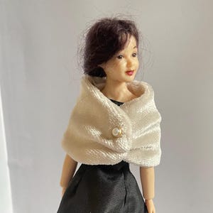 May include: A doll with dark brown hair wearing a white textured velvet shawl fastened with a single pearl-like button over a black satin dress. The doll has a fair complexion, painted facial features, and a neutral expression, presented from the chest up.