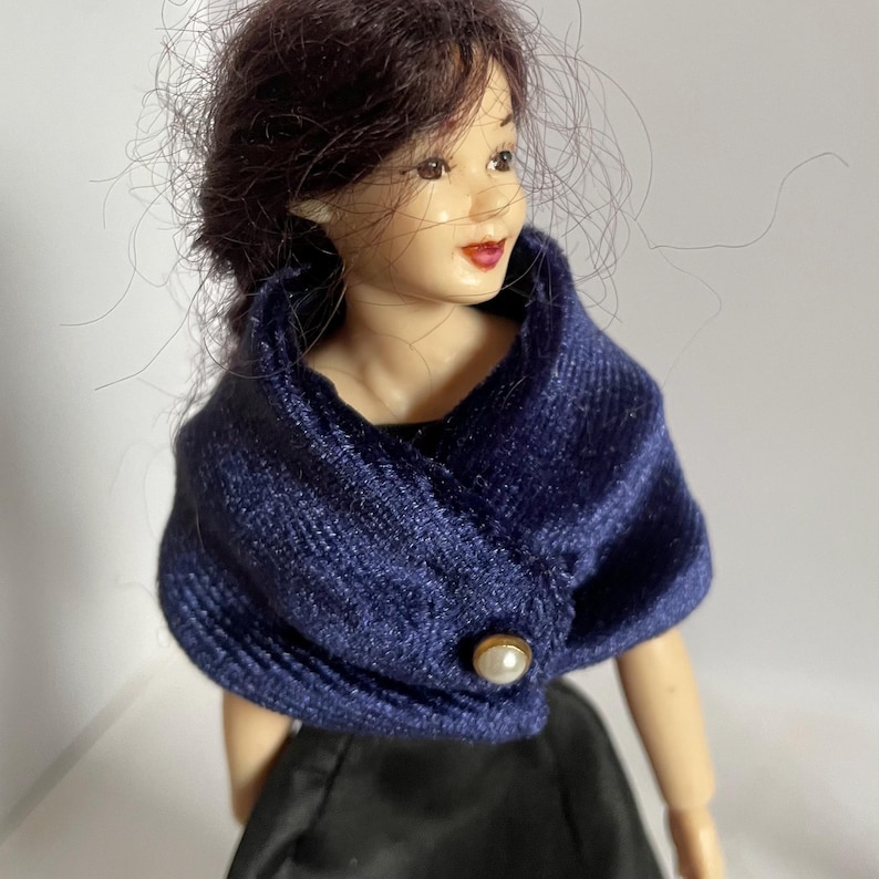 May include: A doll with dark brown hair wearing a navy blue velvet shawl featuring a single white pearl button closure over a black garment. The doll has a light skin tone, painted facial features, and a soft, textured fabric wrap around the shoulders.