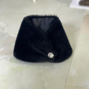 May include: A black faux fur collar featuring a soft, textured pile and a smooth black fabric lining. The collar is secured at the front with a single, clear, faceted crystal-like button that adds a subtle reflective detail to the dark material.