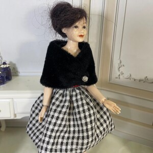 May include: A doll with dark brown hair styled in an updo, wearing a black fuzzy bolero jacket with a single silver button closure over a black and white checkered skirt. The doll has painted facial features including red lips and light skin tone.