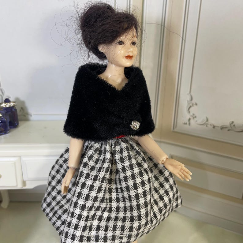 May include: A doll with dark brown hair styled in an updo, wearing a black fuzzy bolero jacket with a single silver button closure over a black and white checkered skirt. The doll has painted facial features including red lips and light skin tone.