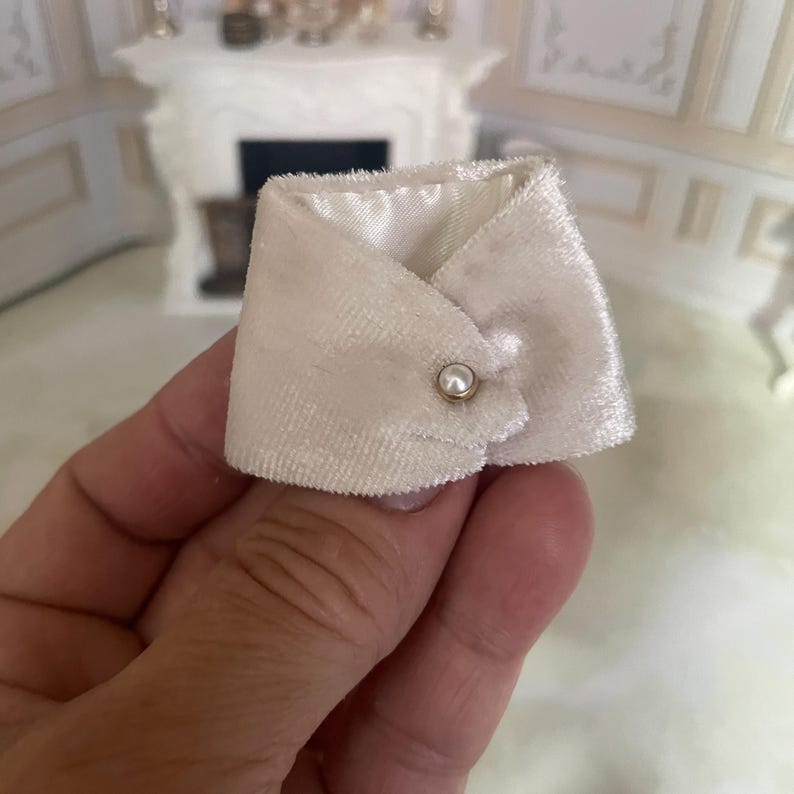 May include: A miniature white velvet collar featuring a pointed design and a single small faux pearl button closure. The collar has a soft, textured surface and a smooth white lining, designed for use as a doll accessory or decorative item.