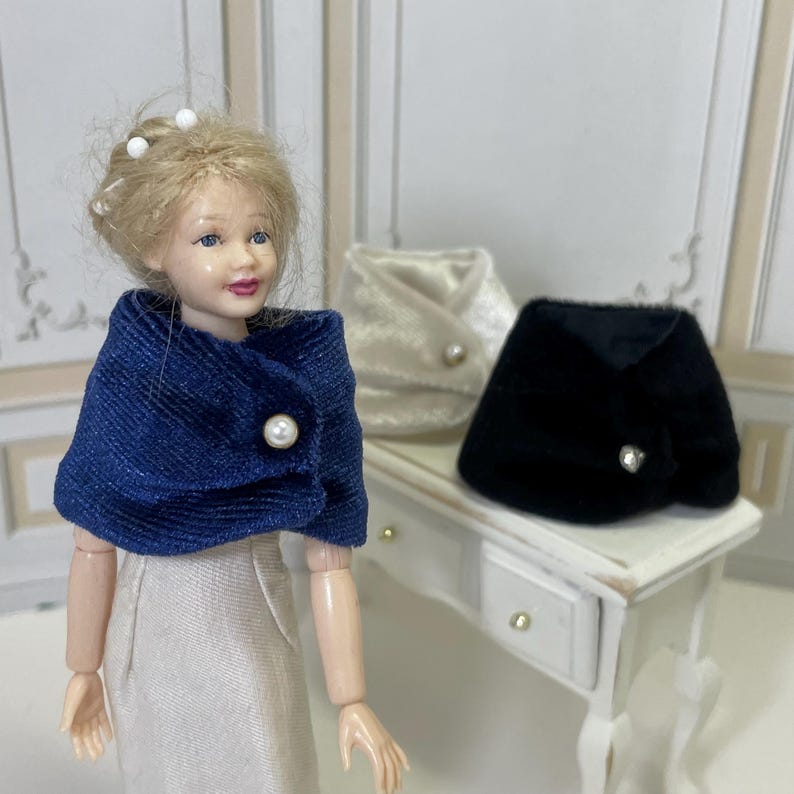 May include: A doll wearing a textured blue capelet with a single white pearl button. Two additional capelets are visible in the background, one in cream and one in black, each featuring a single pearl button closure and a soft, plush fabric texture.