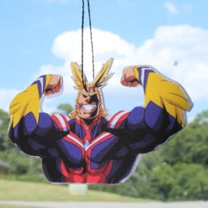 May include: A car air freshener featuring a cartoon character with yellow hair, a red, white, and blue costume, and a determined expression. The character is flexing both arms, showing off their muscles.